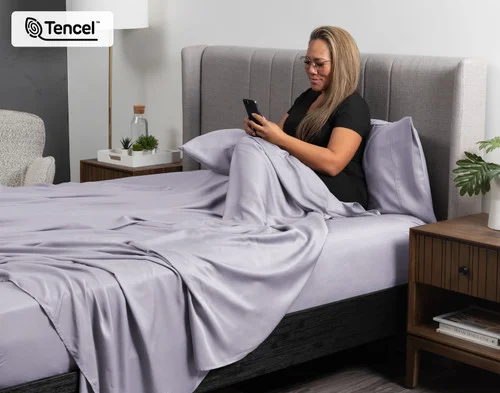 Woman sitting upright in a bed reading her book curled up in the flat sheet of our BeechBliss TENCEL™ Modal Sheet Set in Lavender.