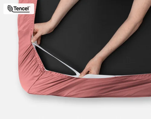 Close-up on two hands stretching the elastic snug fit band on the corner of our Eucalyptus Luxe TENCEL™ Lyocell Fitted Sheet in Raspberry Ice.