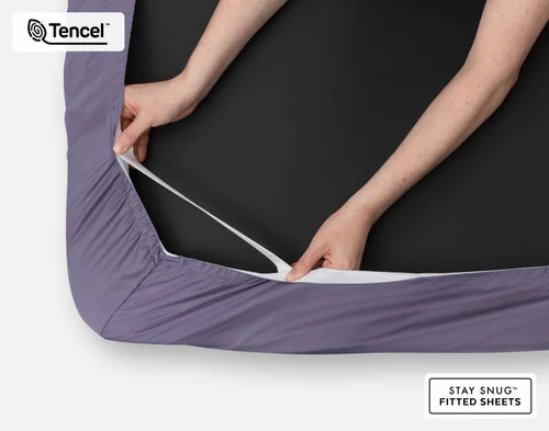Close-up on two hands stretching the elastic snug fit band on the corner of our Eucalyptus Luxe Fitted Sheet in Purple Basil.