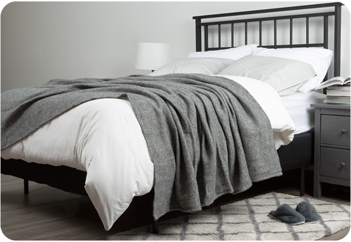 Angled view of a wool blend blanket dressed over a queen sized bed in a white and grey bedroom.
