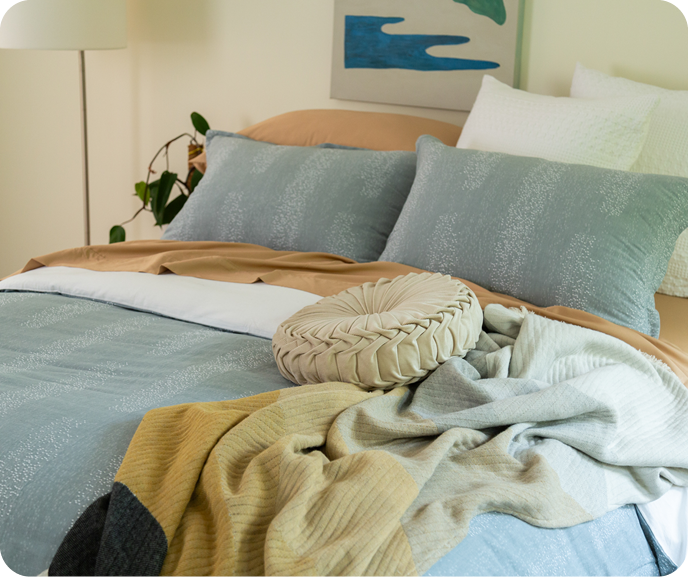 The Aquatico Duvet Cover in blue with a frothy water pattern dressing a bed with brown-orange sheets, the Estrela throw blanket, a cream throw cushion, and white square cushions. The Aquatico Duvet Cover in blue with a frothy water pattern dressing a bed with brown-orange sheets, the Estrela throw blanket, a cream throw cushion, and white square cushions.