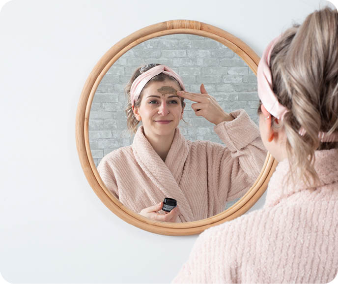 A person applies skincare to their face in front of a round mirror, wearing a pink plush robe and silk headband. A person applies skincare to their face in front of a round mirror, wearing a pink plush robe and silk headband.