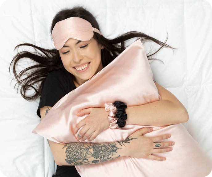 A person lies down hugging one of our Mulberry Silk Pillowcases in Blush, wearing an eye mask in the same fabric and colour.s A person lies down hugging one of our Mulberry Silk Pillowcases in Blush, wearing an eye mask in the same fabric and colour.s