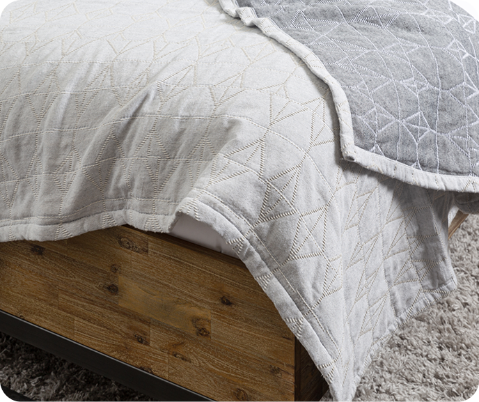Close up of the quilted grey Bragado Blanket from the Portugal Reserve Collection at the corner of a wooden bed with white sheets. Close up of the quilted grey Bragado Blanket from the Portugal Reserve Collection at the corner of a wooden bed with white sheets.