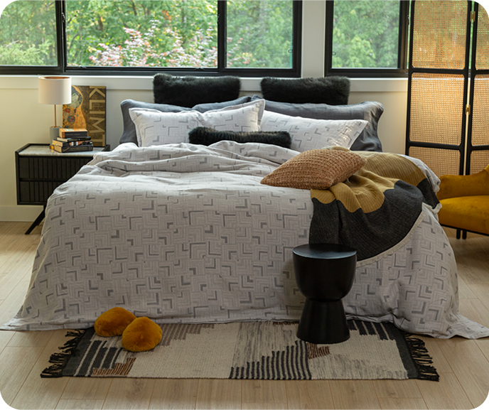 The geometric silver Centro Duvet Cover from the Portugal Reserve Collection with its matching pillow shams paired with black and orange cushions and an orange throw pillow in an artsy bedroom. The geometric silver Centro Duvet Cover from the Portugal Reserve Collection with its matching pillow shams paired with black and orange cushions and an orange throw pillow in an artsy bedroom.