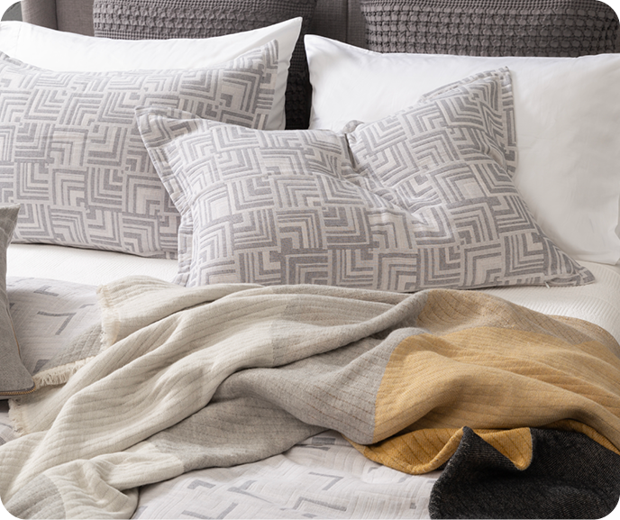 A close up of the head of a bed dressed with the geometric silver Centro Duvet Cover from the Portugal Reserve Collection with its matching pillow shams and the Estrela throw blanket on white sheets. A close up of the head of a bed dressed with the geometric silver Centro Duvet Cover from the Portugal Reserve Collection with its matching pillow shams and the Estrela throw blanket on white sheets.
