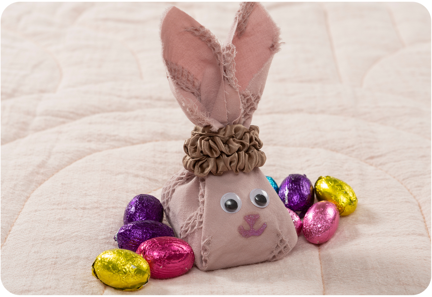 A small cloth DIY bunny treat bag surrounded by eggs, with googly eyes and glitter glue for a face.
