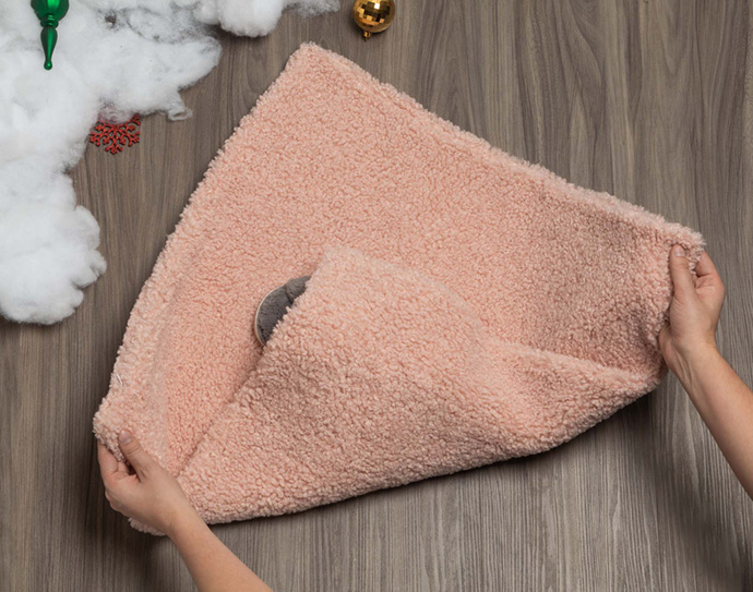 Folding the left and right corners over a folded bottom corner in the center of a fluffy pink pillowsham laying over a wooden surface.