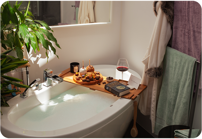A bathtub with a wooden bath caddy covered with an assortment of snacks, a glass of wine and a book. Our Modal cotton towels in Lilac Ash and Seafoam green are are hanging up, alongside our white Plush Faux Fur Bathrobe in Cloud.