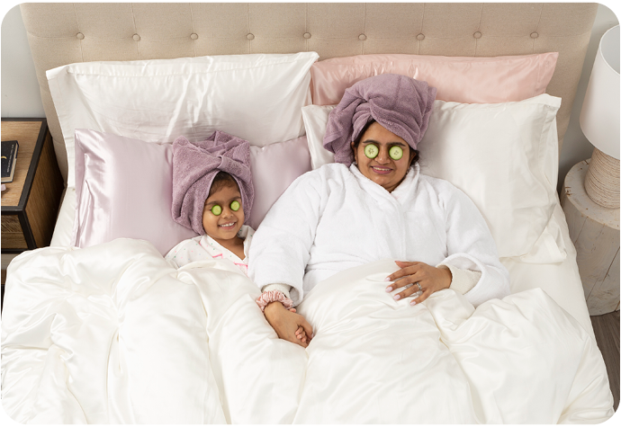 A mother and daughter are pictured laying in bed with cucumber slices covering their eyes, and their hair wrapped in towels. Our Mulberry Silk pillowcases are shown on the white bed.