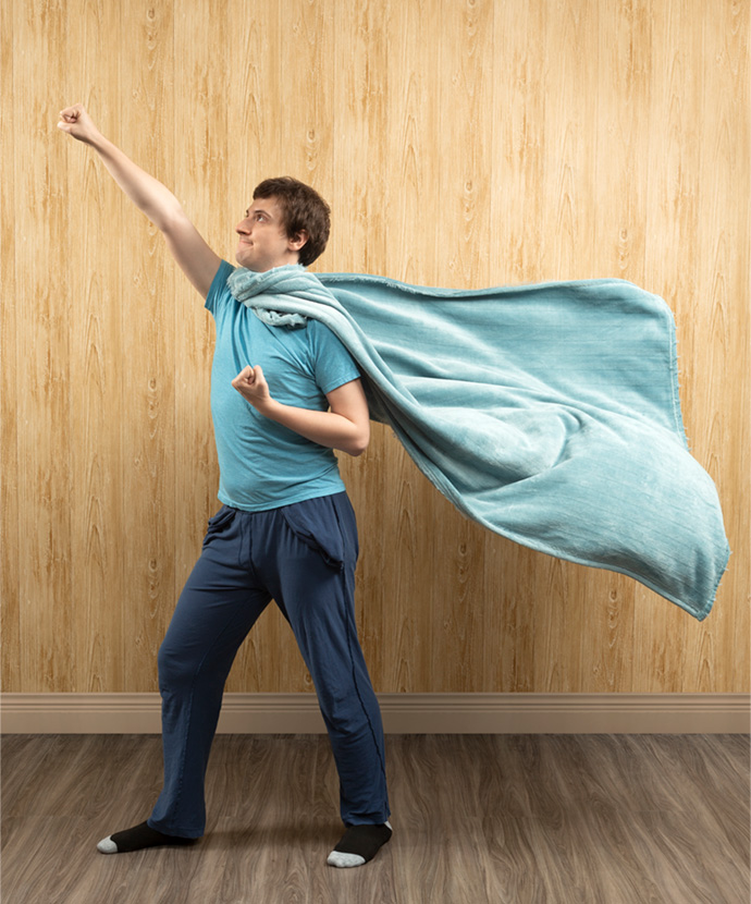 Boy in bright blue pajamas and a soft throw blanket wrapped around his neck as a cape.
