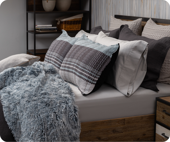 Side view of the Vilanova pillow shams at the head of a bed alongside pillows in different shades of grey and a fluffy blue throw blanket. Side view of the Vilanova pillow shams at the head of a bed alongside pillows in different shades of grey and a fluffy blue throw blanket.